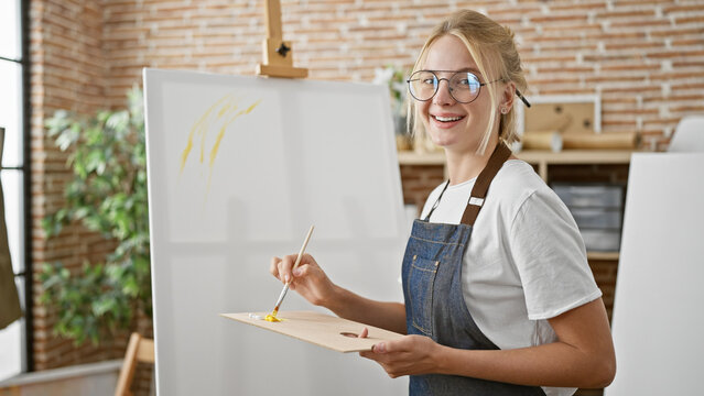 Young blonde woman artist smiling confident holding paintbrush and palette at art studio