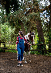 beautiful smiling brunette cowboy woman in red checkered shirt walking with horse in green forest on sunny day