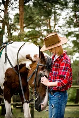Foto op Canvas little handsome blonde smiling  boy  in red checkered shirt and cowboy hat hugging  horse in green forest on sunny day  © Tetatet