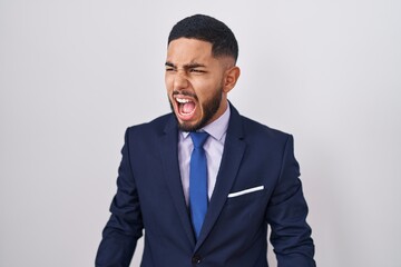 Young hispanic man wearing business suit and tie angry and mad screaming frustrated and furious,...