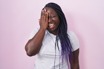 Young african woman standing over pink background covering one eye with hand, confident smile on face and surprise emotion.