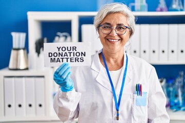 Middle age woman with grey hair working at scientist laboratory holding your donation matters...