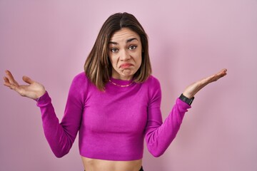 Hispanic woman standing over pink background clueless and confused expression with arms and hands...