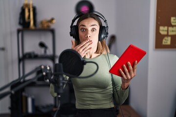 Hispanic woman singing song using microphone and tablet covering mouth with hand, shocked and...