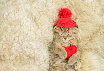 Cute fold tabby kitten wearing warm hat hugs red heart on the bed at home. Valentines day concept. Top down view. Empty space for text