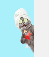 Happy cat with towel on it head and with  pieces of cucumber on it eyes holds tropic cocktail behind empty white banner. isolated on blue background