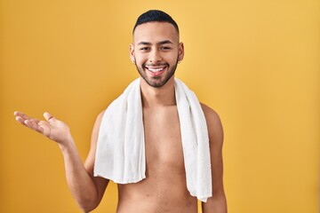 Young hispanic man standing shirtless with towel smiling cheerful presenting and pointing with palm of hand looking at the camera.