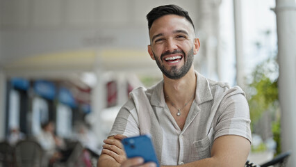 Young hispanic man using smartphone sitting on table smiling at coffee shop terrace
