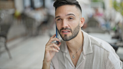 Young hispanic man talking on smartphone sitting on table at coffee shop terrace