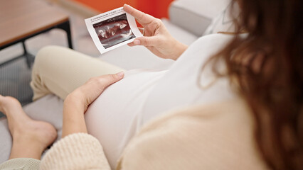 Young pregnant woman sitting on sofa looking baby ultrasound at home