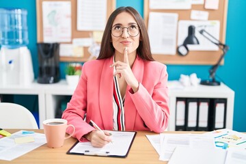 Young hispanic woman working at the office wearing glasses thinking concentrated about doubt with...