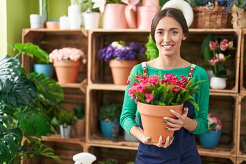 Young hispanic woman working at florist shop holding plant looking positive and happy standing and smiling with a confident smile showing teeth