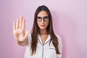 Young brunette woman wearing glasses standing over pink background doing stop sing with palm of the...