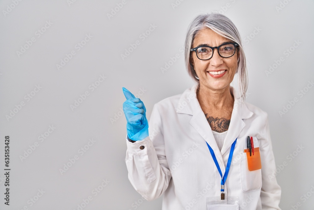 Wall mural middle age woman with grey hair wearing scientist robe doing happy thumbs up gesture with hand. appr