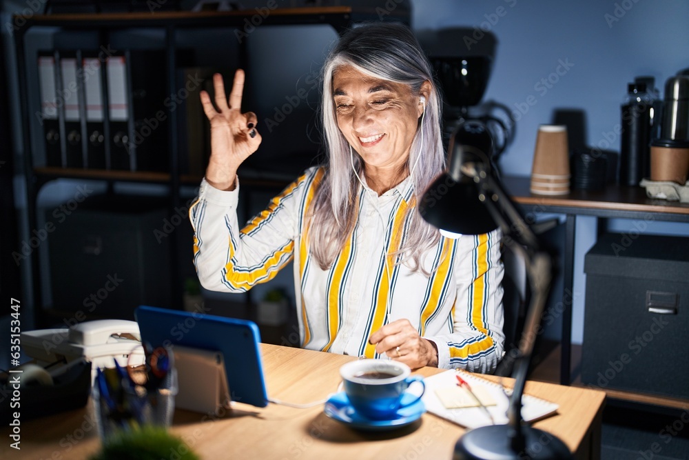 Wall mural middle age woman with grey hair working at the office at night smiling positive doing ok sign with h