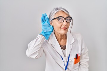 Middle age woman with grey hair wearing scientist robe smiling with hand over ear listening an hearing to rumor or gossip. deafness concept.