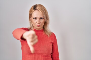 Blonde woman standing over isolated background looking unhappy and angry showing rejection and negative with thumbs down gesture. bad expression.