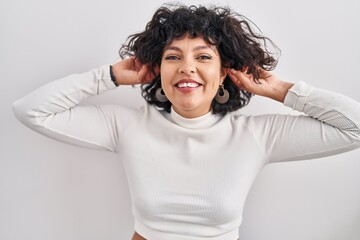 Hispanic woman with curly hair standing over isolated background smiling pulling ears with fingers,...