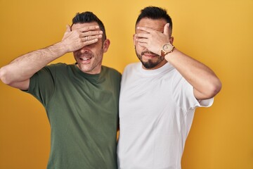 Homosexual couple standing over yellow background smiling and laughing with hand on face covering eyes for surprise. blind concept.