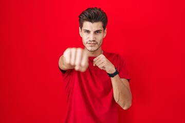 Young hispanic man standing over red background punching fist to fight, aggressive and angry...