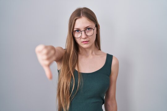 Young caucasian woman standing over white background looking unhappy and angry showing rejection and negative with thumbs down gesture. bad expression.