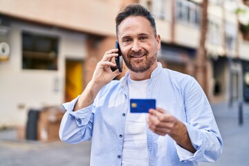 Young caucasian man talking on smartphone holding credit card at street