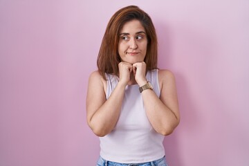 Brunette woman standing over pink background laughing nervous and excited with hands on chin...