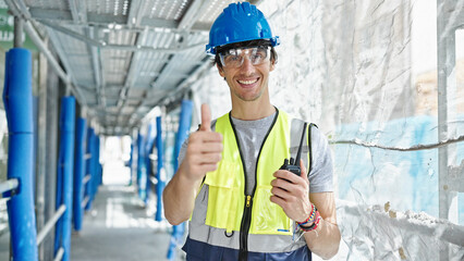 Young hispanic man architect holding walkie-talkie doing thumb up gesture at construction place
