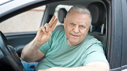 Middle age grey-haired man smiling confident sitting on car doing victory gesture at street