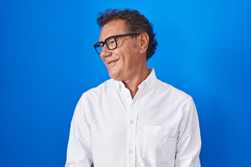 Middle age hispanic man standing over blue background looking away to side with smile on face, natural expression. laughing confident.