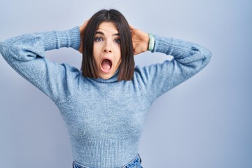 Young hispanic woman standing over blue background crazy and scared with hands on head, afraid and...