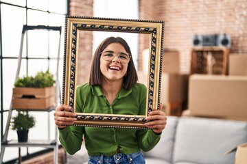 Young hispanic woman at new home holding empty frame smiling and laughing hard out loud because funny crazy joke.