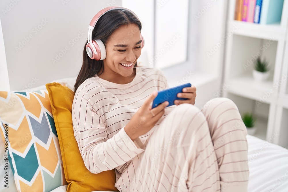 Sticker young beautiful hispanic woman playing video game sitting on bed at street