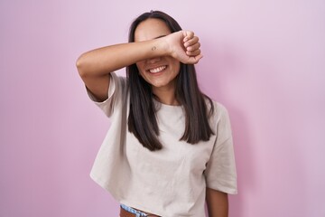 Fototapeta premium Young hispanic woman standing over pink background covering eyes with arm smiling cheerful and funny. blind concept.