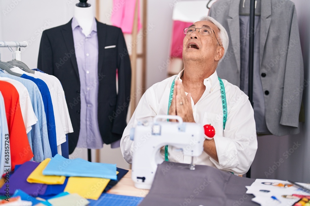 Poster middle age man with grey hair dressmaker using sewing machine begging and praying with hands togethe