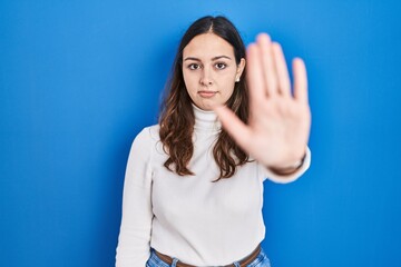 Young hispanic woman standing over blue background doing stop sing with palm of the hand. warning...