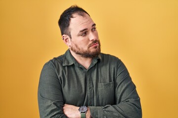 Plus size hispanic man with beard standing over yellow background looking to the side with arms crossed convinced and confident