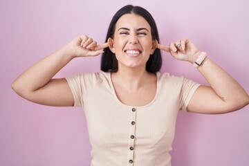 Young hispanic woman standing over pink background smiling pulling ears with fingers, funny...