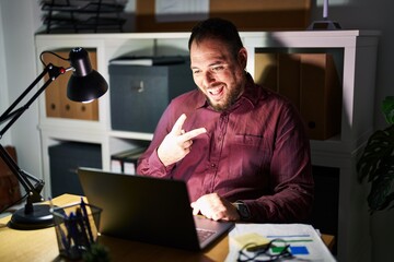 Plus size hispanic man with beard working at the office at night smiling with happy face winking at the camera doing victory sign. number two.