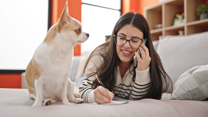 Young hispanic woman with chihuahua dog speaking on the phone and taking notes lying on the sofa at home