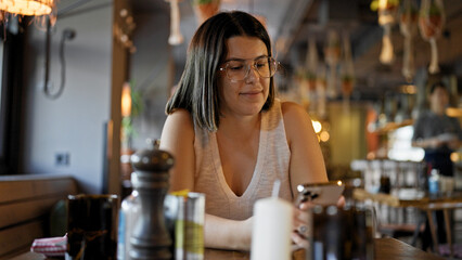Young beautiful hispanic woman using smartphone sitting on the table at the restaurant