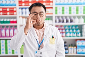Chinese young man working at pharmacy drugstore mouth and lips shut as zip with fingers. secret and...