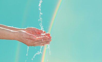 Water pouring on female hands in sunlight. Hands with water stream on rainbow background for eco concept of water scarcity or Earth Day.