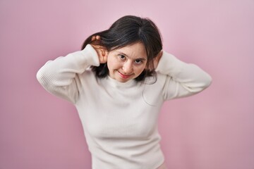 Woman with down syndrome standing over pink background trying to hear both hands on ear gesture, curious for gossip. hearing problem, deaf