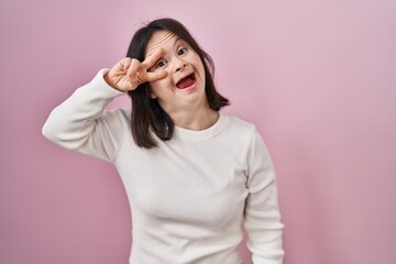 Woman with down syndrome standing over pink background doing peace symbol with fingers over face,...