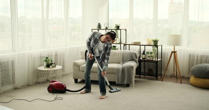 Crazy young man wearing headphones, who is cleaning the living room and dancing simultaneously. With dance moves and a lively spirit, he brings a unique and entertaining flair to the cleaning process.