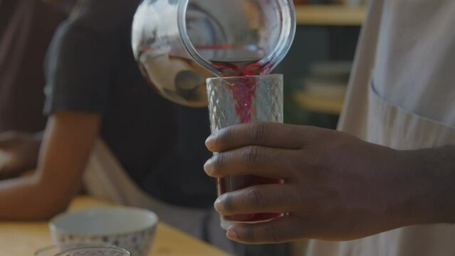 Close up shot of hands of unrecognizable black man pouring red Zobo tea into glass while cooking traditional meal with family in kitchen at home