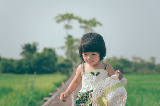 Portrait photo of a cute infant girl is costumed in green dress and holding white hat during walking of wooden bridge with background of outdoor rice field in sunny day.