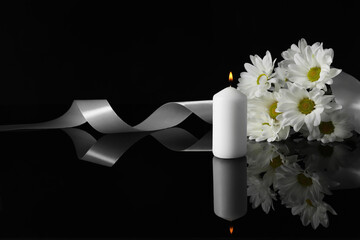 Burning candle, white chrysanthemum flowers and ribbon on black mirror surface in darkness, space...