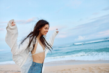 Brunette woman with long hair in a white shirt and denim shorts smile and happiness running on the beach and having fun smile with teeth in front of the ocean, vacation summer trip